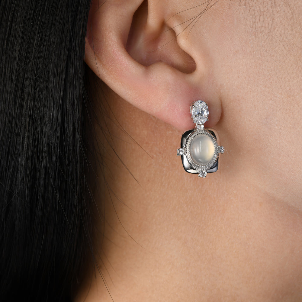 Experience refined elegance with our earrings, each featuring an oval white chalcedony gracefully nestled in the heart of a rounded rectangle platinum base. Adorned with four small round simulated diamonds, this piece exudes timeless sophistication, and it descends alongside a smaller oval white simulated diamond for a touch of graceful charm.