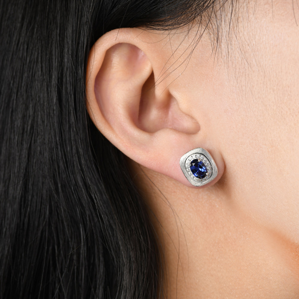 These stud earrings featuring an enchanting oval sapphire encircled by glistening white simulated diamonds. The centerpiece is gracefully nestled on a cushion-shaped platinum base intricately processed with engraving techniques.